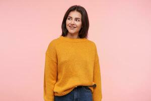 allegro giovane attraente Marrone dai capelli donna con casuale acconciatura sorridente piacevolmente e conservazione mani lungo corpo mentre in piedi al di sopra di rosa sfondo foto