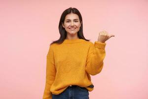 affascinante giovane allegro Marrone dai capelli femmina con perdere capelli conservazione sua mani sollevato mentre mostrando a parte e sorridente piacevolmente, in piedi al di sopra di rosa sfondo foto