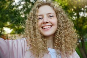 selfie-ritratto di affascinante giovane bellissimo bionda ragazza con Riccio capelli, nel un' grande umore, bene sorridente, al di sopra di natura sfondo foto