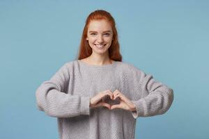 romantico ritratto di tenero affascinante donna su un' blu sfondo. ragazza con rosso capelli Spettacoli cuore forma gesto con mani. femmina con treccia nel camicia mostrando amore simbolo, guardare a telecamera foto