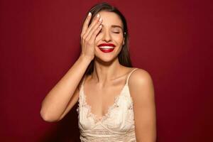 lieta giovane attraente buio dai capelli donna con rosso labbra conservazione sollevato palma su sua viso e sorridente felicemente con chiuso occhi, in posa al di sopra di Borgogna sfondo foto