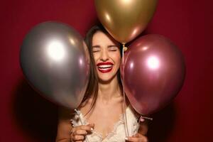 piacevole guardare giovane gioioso buio dai capelli elegante signora conservazione sua occhi chiuso e ridendo felicemente mentre avendo festa con sua gli amici, in posa al di sopra di Borgogna sfondo foto