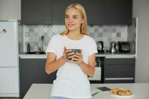 ritratto di contento giovane bionda ragazza nel un' bene umore sta Leans su il tavolo, sorridente, avendo colazione, mantiene un' grande grigio tazza da tè, guardare lontano, sognando, su un' cucina sfondo foto