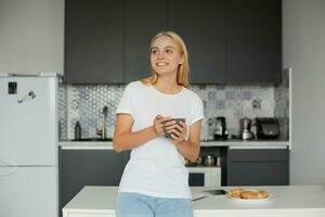 contento giovane bionda ragazza nel un' bene umore sta Leans su il tavolo, sorridente, avendo colazione, mantiene un' grande grigio tazza da tè, guardare lontano, sognando, pensiero di il In arrivo giorno, su un' cucina sfondo foto