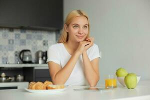 foto di giovane bionda ragazza si siede a il cucina tavolo nel il mattina, sorridente, sembra lieto per il finestra, su il tavolo -un bicchiere di succo, mele, panini, vestito nel un' domestico maglietta, cucina sfondo