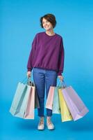 interno foto di bene guardare giovane lieto buio dai capelli Riccio donna vestito nel casuale Abiti sorridente piacevolmente mentre in piedi al di sopra di blu sfondo con sua acquisti