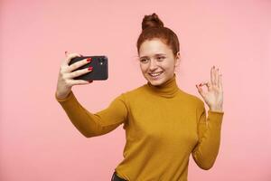 lieto giovane bellissimo testa Rossa donna con casuale acconciatura raccolta mano con ok gesto mentre fabbricazione autoscatto con sua mobile Telefono, in piedi contro rosa sfondo foto