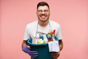 allegro giovane bene guardare brunetta maschio nel occhiali conservazione il suo occhi chiuso e sorridente felicemente mentre Tenere pulizia roba, isolato al di sopra di rosa sfondo foto