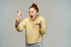 arrabbiato guardare donna, urlando testa Rossa ragazza con capelli panino. indossare giallo maglione e Tenere un' smartphone. discutere con qualcuno su video Chiacchierare. Incazzata spento. In piedi isolato al di sopra di grigio sfondo foto