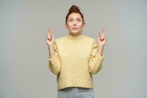 ritratto di attraente, adulto testa Rossa ragazza con capelli si riunirono nel un' panino. indossare pastello giallo maglione e jeans. attraversare dita, fabbricazione un' desiderio. Guardando su a copia spazio, isolato al di sopra di grigio sfondo foto