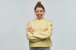 adolescenziale ragazza, contento guardare donna con biondo capelli si riunirono nel panino. indossare giallo maglione. mantiene braccia attraversato e a grandi linee sorridente. occhiolino a il telecamera, isolato al di sopra di bianca sfondo foto