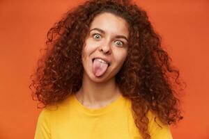 vicino su. simpatico guardare donna, bellissimo ragazza con Riccio Zenzero capelli. indossare giallo maglietta. persone e emozione concetto. mostrando sua lingua. Guardando a il telecamera, isolato al di sopra di arancia sfondo foto