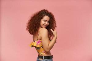 timido guardare donna, bellissimo ragazza con Riccio Zenzero capelli. indossare colorato senza spalle camicetta. persone e emozione concetto. Guardando a il telecamera al di sopra di spalla, isolato al di sopra di pastello rosa sfondo foto