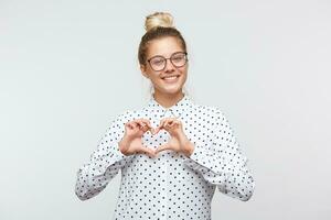 romantico ritratto di tenero affascinante donna su un' bianca sfondo. ragazza Spettacoli cuore forma gesto con mani. femmina nel un' bianca camicia con nero punti, occhiali, mostrando amore simbolo, guardare a telecamera foto