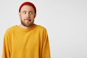 ritratto di un' uomo con un' rosso barba, indossare casuale maglione e un' rosso cappello, vigliaccamente, con cautela inseguendo il suo labbro, sguardi lato destro a il vuoto spazio per copia, isolato su bianca sfondo foto