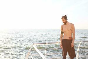 giovane sportivo tipo con barba, riposo dopo mattina jogging a il mare, ascolta preferito mescolare su cuffia. foto