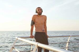 ritratto di giovane sportivo uomo con barba, riposo dopo jogging a il mare, ascolta preferito canzone su cuffia. gode greates mattina foto