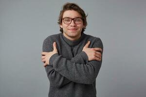 ritratto di giovane sorridente uomo con bicchieri indossa nel grigio maglione, sta al di sopra di grigio sfondo e abbracci lui stesso, sembra felicità. foto