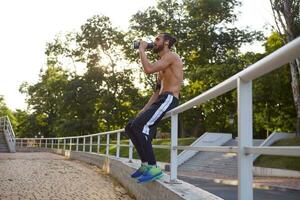 giovane attraente barbuto tipo ha estremo sport nel parco, riposo a il parco dopo jogging, potabile acqua, conduce salutare attivo stile di vita. fitness maschio modello. foto