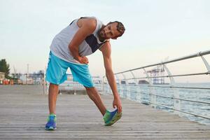 foto di giovane sportivo barbuto tipo fare allungamento, mattina esercizi di il mare, riscaldamento dopo correre, conduce salutare attivo stile di vita. fitness e salutare concetto.