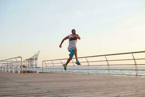 foto di giovane sportivo bello barbuto in esecuzione tipo fare mattina esercizi di il mare, riscaldamento prima correre, conduce salutare attivo stile di vita. fitness e sport concetto.