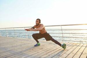 giovane maschile sportivo barbuto tipo fare allungamento, ascoltando preferito musica su cuffia, mattina esercizi di il mare, riscaldamento dopo correre, conduce salutare attivo stile di vita. fitness maschio modello. foto