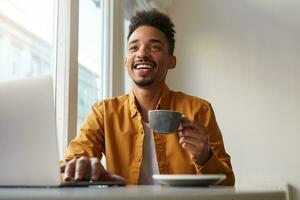 giovane buio spellato sorridente tipo seduta nel un' bar e Lavorando su un' il computer portatile e Guardando un' divertente video, indossa nel giallo camicia, bevande aromatico caffè , sorridente e gode il opera di un' libero professionista. foto