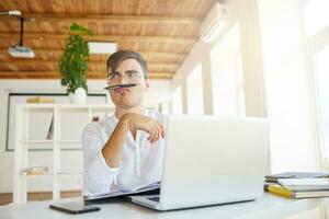 avvicinamento di divertente pensieroso giovane uomo d'affari indossa bianca camicia e spettacoli con il computer portatile e cellula Telefono pensiero e fabbricazione divertente viso a il tavolo nel ufficio foto