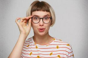 avvicinamento di contento sorpreso giovane donna indossa a strisce t camicia e spettacoli si sente stupito e sorridente isolato al di sopra di bianca sfondo foto