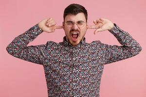 vicino su di urlando urlando arrabbiato giovane barbuto uomo vestito nel colorato camicia, mantiene chiuso occhi, Due dita vicino il suo orecchie, mostrando gesto di sordità, ignora qualcuno, al di sopra di rosa parete foto