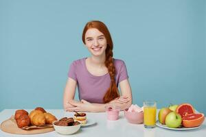 sorridente dai capelli rossi ragazza con intrecciato capelli seduta a un' tavolo, di per mangiare prima colazione guardare a il telecamera, isolato su blu parete. su il tavolo posare cottura al forno e fresco frutta succo Yogurt. cibo concetto foto