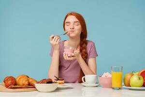 simpatico dai capelli rossi ragazza provando degustazione ciliegia Yogurt wirh un' cucchiaino, chiuso sua occhi a partire dal piacere, seduta a il tavolo durante il pranzo, pasticcini su il tavolo e fresco frutta, contro un' blu sfondo. foto