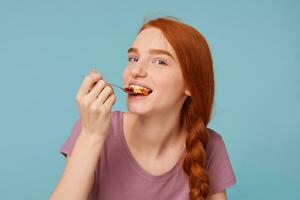 un' simpatico e attraente dai capelli rossi ragazza cerca degustazione mangia qualcosa delizioso, sembra in il telecamera scherzosamente mette un' cucchiaio con muesli nel sua bocca, isolato su un' blu sfondo. foto