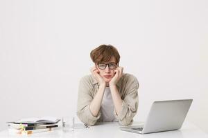 avvicinamento di infelice stanco giovane uomo alunno indossa bicchieri e beige camicia sembra triste e Lavorando con il computer portatile computer e i Quaderni a il tavolo isolato al di sopra di bianca sfondo foto