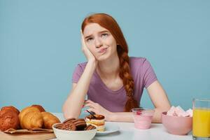 studio foto di triste dai capelli rossi ragazza,dubbi medita pensiero di cibo, salute, dieta, extra calorie, cottura al forno prodotti e fresco frutta succo Yogurt siamo su il tavolo, isolato su un' blu sfondo