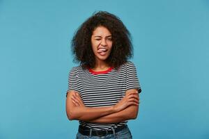 studio tiro di divertente africano americano donna Spettacoli lingua, ha giocoso espressione, afro acconciatura, fa smorfia, vestito nel nudo maglietta, isolato al di sopra di blu sfondo. comico buio spellato ragazza foto