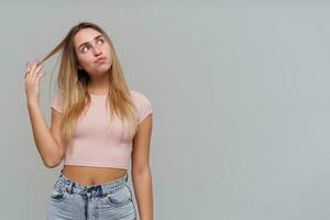 pensiero ragazza, riflessivo guardare donna con biondo lungo capelli. indossare rosa maglietta e jeans. giocando con filo di capelli. Guardando per il giusto a copia spazio, isolato al di sopra di grigio sfondo foto