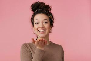 adolescenziale ragazza, contento guardare donna con buio Riccio capelli panino. indossare fascia per capelli, orecchini e Marrone maglione. ha rendere su. sorridente al di sopra di palma. Guardando a il telecamera isolato al di sopra di pastello rosa sfondo foto