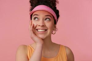 vicino su di sognare donna con buio Riccio capelli panino. indossare rosa visiera, orecchini, arancia serbatoio superiore. detiene palma sotto sua mento. Guardando per il sinistra a copia spazio, isolato al di sopra di rosa sfondo foto