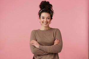 carino guardare donna, bellissimo ragazza con buio Riccio capelli panino. indossare fascia per capelli, orecchini e Marrone maglione. ha rendere su. detiene braccia incrociato. Guardando a il telecamera isolato al di sopra di pastello rosa sfondo foto