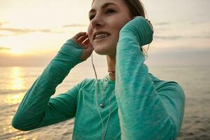 giovane a grandi linee sorridente donna a il mare nel il mattina, ascoltando preferito musica a cuffia. Salute cura concetto. foto