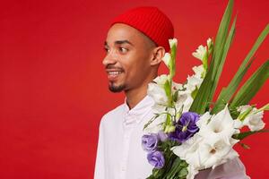 vicino su di giovane positivo buio spellato uomo, indossa nel bianca camicia e rosso cappello, sta al di sopra di rosso sfondo e a grandi linee sorridente. foto