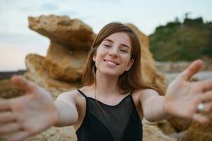 vicino su di giovane allegro Zenzero carino signora con lentiggini su il spiaggia, traino mani per il telecamera, a grandi linee sorridente e sembra positivo e contento. foto