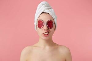 vicino su di triste giovane donna con un' asciugamano su sua testa dopo doccia, con maschera su occhi, sembra disconnesso, sta al di sopra di rosa sfondo. foto