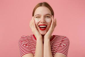 carino giovane contento dai capelli rossi signora nel un' rosso a strisce maglietta, con rosso labbra , molto lieta con nuovo cerotti, occhiolino e a grandi linee sorridente, tocchi il suo viso con dita. sta al di sopra di rosa sfondo. foto