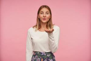 ritratto di affascinante giovane femmina con bionda capelli raccolta palma e soffiaggio aria bacio per telecamera, essere nel simpatico umore, indossare casuale Abiti mentre in piedi al di sopra di rosa sfondo foto