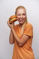 divertente tiro di bello giovane bionda donna con coda di cavallo acconciatura dando occhiolino per telecamera e mostrando sua lingua mentre in posa al di sopra di bianca sfondo con fresco hamburger nel sua mani foto