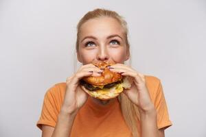 ritratto di lieta giovane bello bionda donna con casuale acconciatura mangiare fresco Hamburger con grande appetito e guardare allegramente verso l'alto, in posa al di sopra di bianca sfondo foto