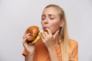 ritratto di giovane soddisfatto bionda femmina con casuale acconciatura degustazione sua fresco Hamburger con grande piacere e conservazione occhi Chiuso, in piedi contro bianca sfondo foto