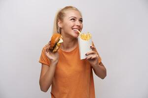 interno foto di attraente giovane bionda signora nel arancia maglietta sorridente allegramente mentre traino con denti francese patatine fritte e conservazione Hamburger nel altro mano, isolato al di sopra di bianca sfondo
